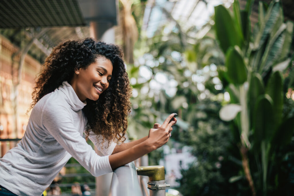 A curly woman smiling while using her phone with the outdoors as her background